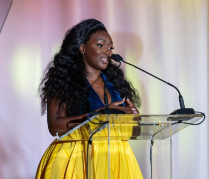 Woman with dark curly hair wearing blue and yellow dress speaks at podium