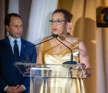 Woman with short hair and yellow dress wearing glasses speaks at podium with man and woman behind her