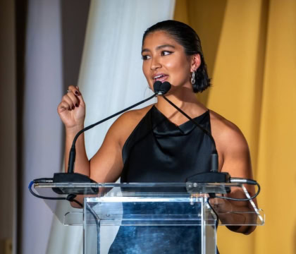 Woman with short dark hair and black dress speaks at podium with arm extended