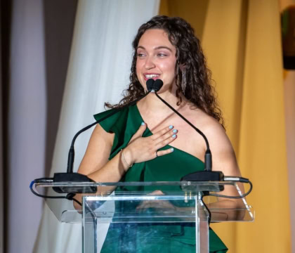 Woman with brown curly hair wearing green dress speaks at podium with hand over heart