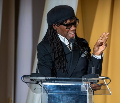 Man with long hair wearing suit, hat, and sunglasses speaks at podium while gesturing off camera