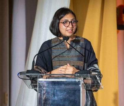 Woman with dark hair and long dark top wearing glasses speaks at podiums with hands together