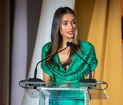 Woman with straight, dark-blonde hair wearing flowy green dress speaks at podium