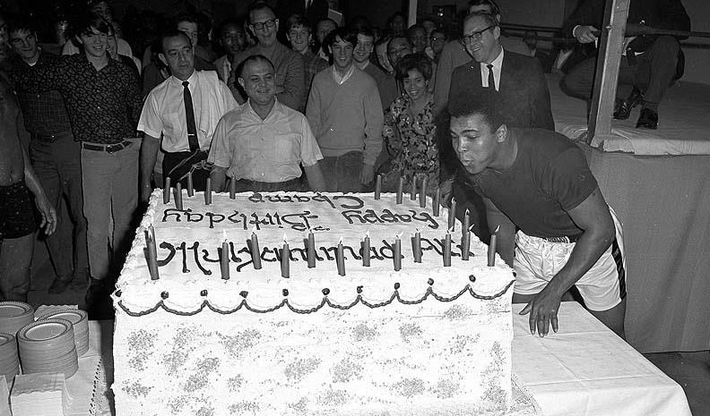 Black and white photo of Muhammad Ali blowing out candles on big birthday cake