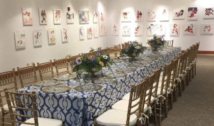 A long banquet table set with chairs, chargers, and floral arrangements in an art gallery