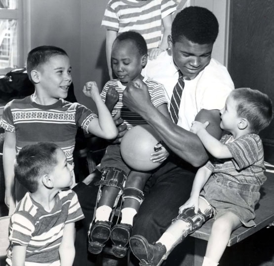 Black and white photo of Muhammad Ali in a shirt and tie with a number of young children in leg braces