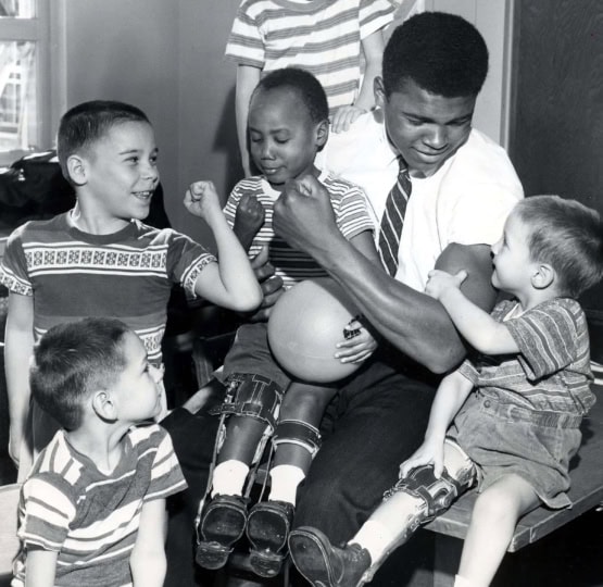 Black and white photo of Muhammad Ali in a shirt and tie with a number of young children in leg braces