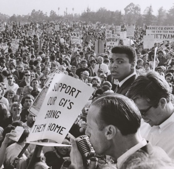 Black and white photo of Muhammad Ali standing among anti-war protesters holding signs