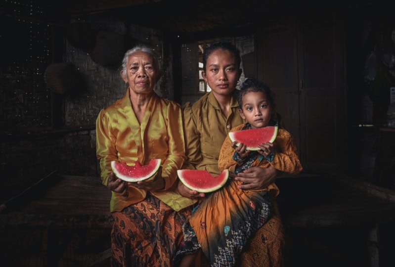 Multiple generations of women sitting and wearing gold outfits, holding onto slices of watermelon and looking directly at camera