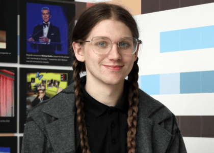 Picture of individual with long. brown, tied-up hair wearing glasses and black shirt/jacket smiling at camera