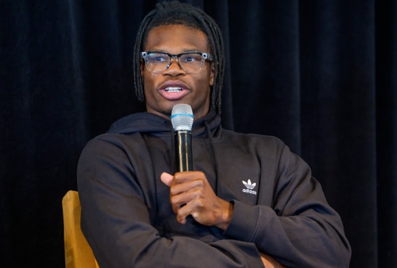 Travis Hunter, wearing glasses and black hoodie, holds microphone while speaking to audience