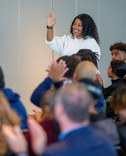 Woman standing in audience wearing white shirt holds hand up for a question