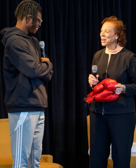 Lonnie Ali holding up boxing gloves and smiling attempting to hand them to Travis Hunter