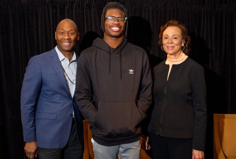 Group photo of man DeVone Holt wearing blue suit, Lonnie Ali wearing black shirt and pants, and Travis Hunter wearing black hoodie and jeans