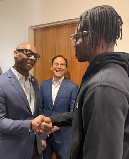 Man in blue suit shakes hand of man in gray hoodie while onlookers smile