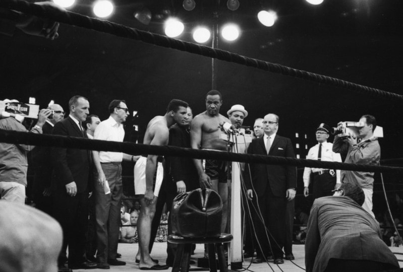Black and white photo of Sonny Liston weighing in around group of people with Muhammad Ali observing