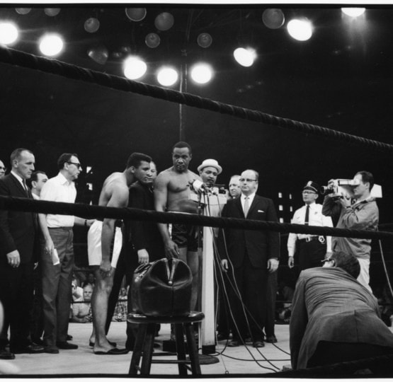 Black and white photo of Sonny Liston weighing in around group of people with Muhammad Ali observing