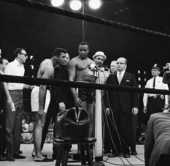 Black and white photo of Sonny Liston weighing in around group of people with Muhammad Ali observing