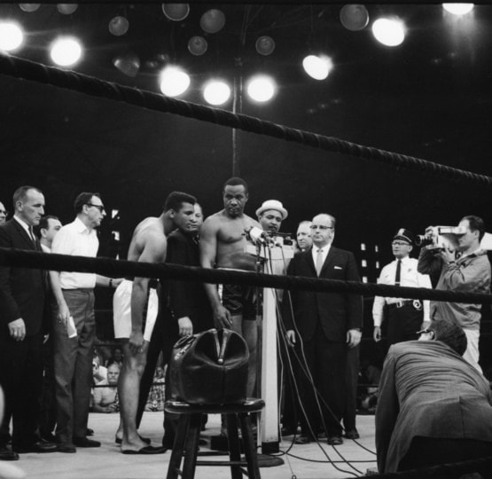 Black and white photo of Sonny Liston weighing in around group of people with Muhammad Ali observing