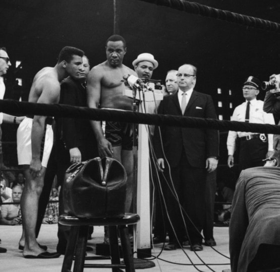 Black and white photo of Sonny Liston weighing in around group of people with Muhammad Ali observing