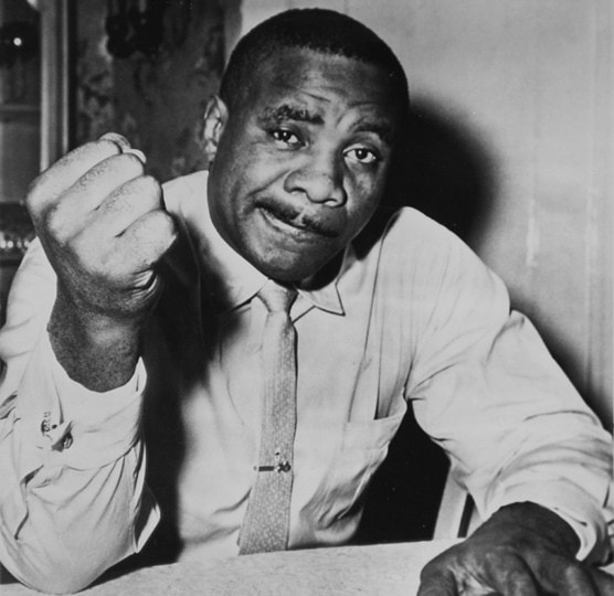Black and white photo of Sonny Liston in button up shirt and tie with right fist extended to camera
