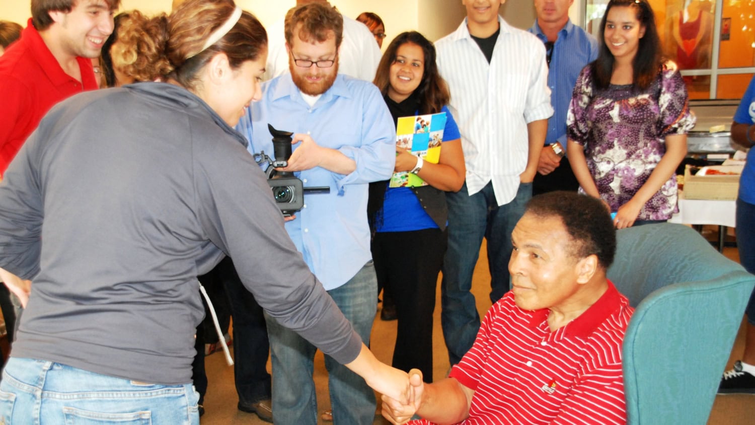 Photo of Muhammad Ali wearing red button down shirt sitting in chair and shaking hands with guests
