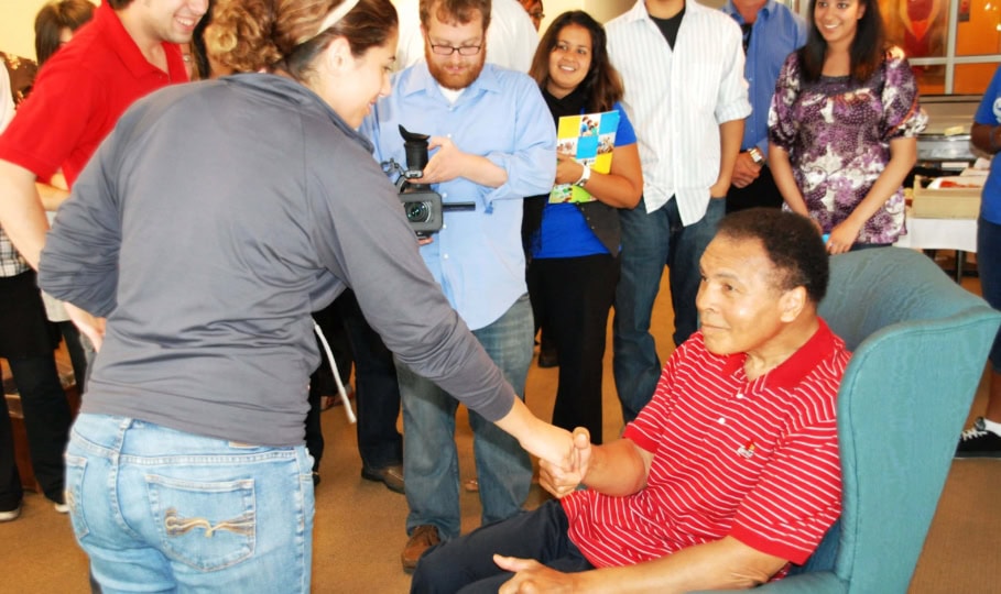 Photo of Muhammad Ali wearing red button down shirt sitting in chair and shaking hands with guests
