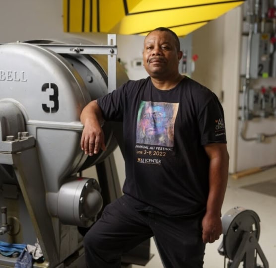 Man wearing black shirt with Ali Fest design rests arms near machinery and poses for camera