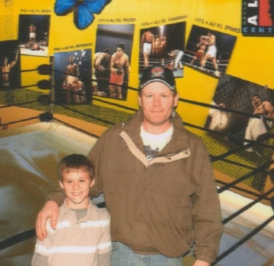 Man in brown jacket and black hat stands with his son and poses for camera in front of boxing ring