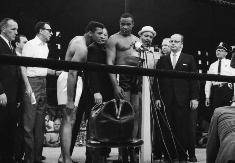 Black and white photo of Sonny Liston weighing in around group of people with Muhammad Ali observing