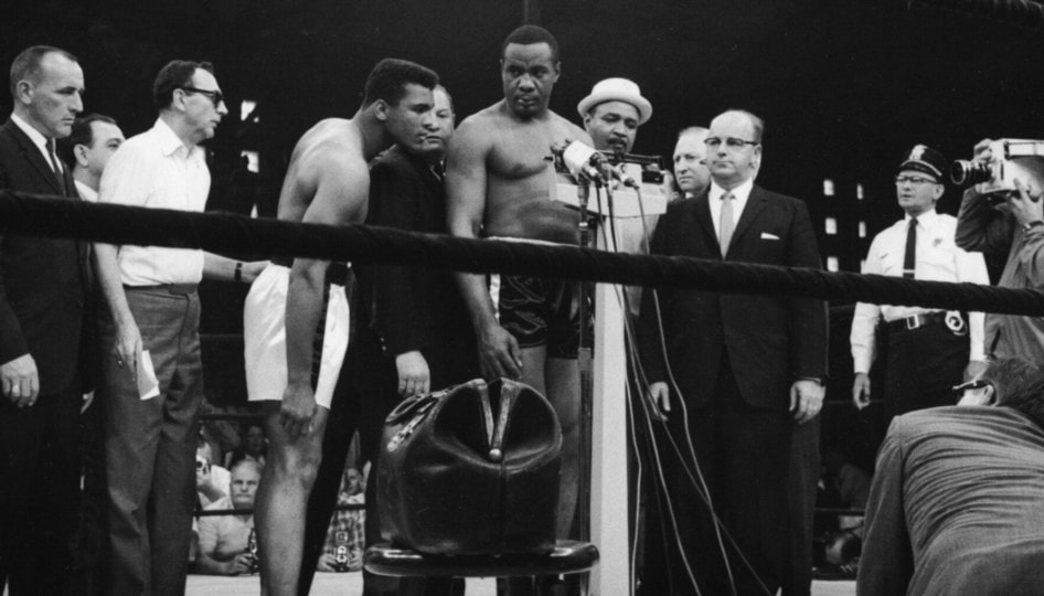 Black and white photo of Sonny Liston weighing in around group of people with Muhammad Ali observing