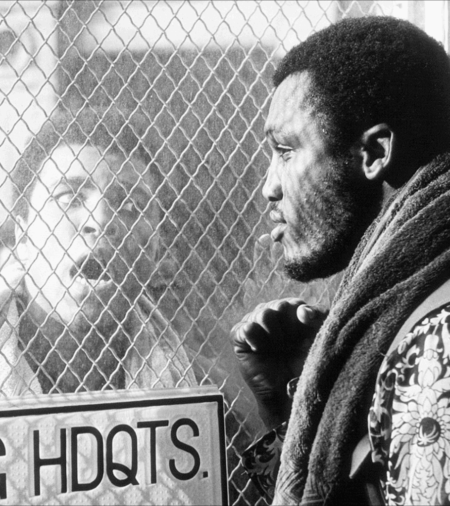 Black and white phot of Muhammad Ali and Joe Frazier looking at each other through opposite sides of a fence