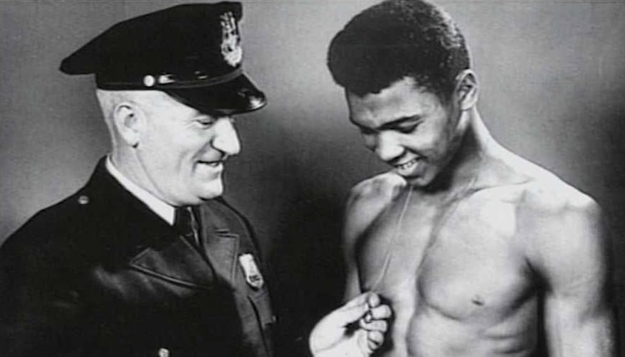 Black and white photo of a young Muhammad Ali shirtless in boxing shorts with Joe Martin in a police uniform