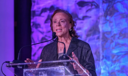 Lonnie Ali speaking on a stage while standing at a clear glass podium in front of a purple background