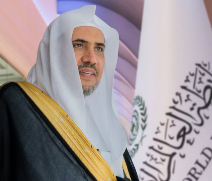 Man dressed in traditional Muslim attire standing in front of flag and looking off camera with arms crossed