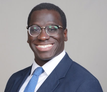 Man with glasses, dark blue suit and light blue tie smiling at camera