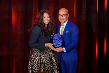 Woman with short brown hair wearing dark blue dress and man with dark glasses and light blue suit hold award and smile at camera