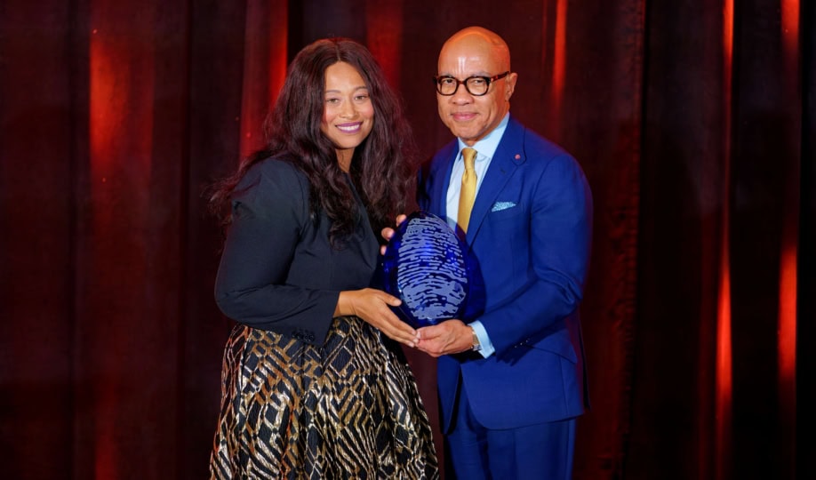 Woman with short brown hair wearing dark blue dress and man with dark glasses and light blue suit hold award and smile at camera