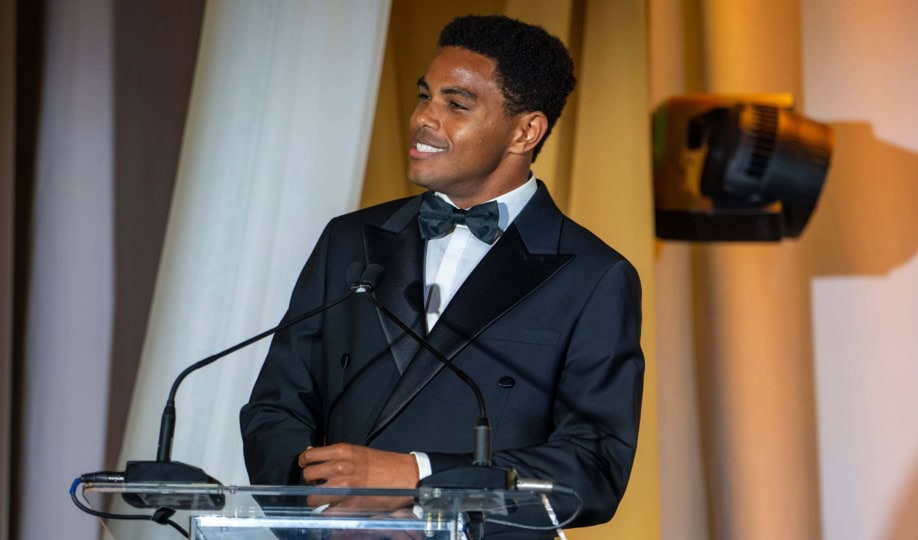 Man wearing black tuxedo and black bow tie standing at podium and smiling