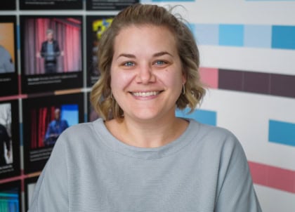 Woman with short, blond, curly hair wearing a gray sweater in front of colorful wall smiling at camera