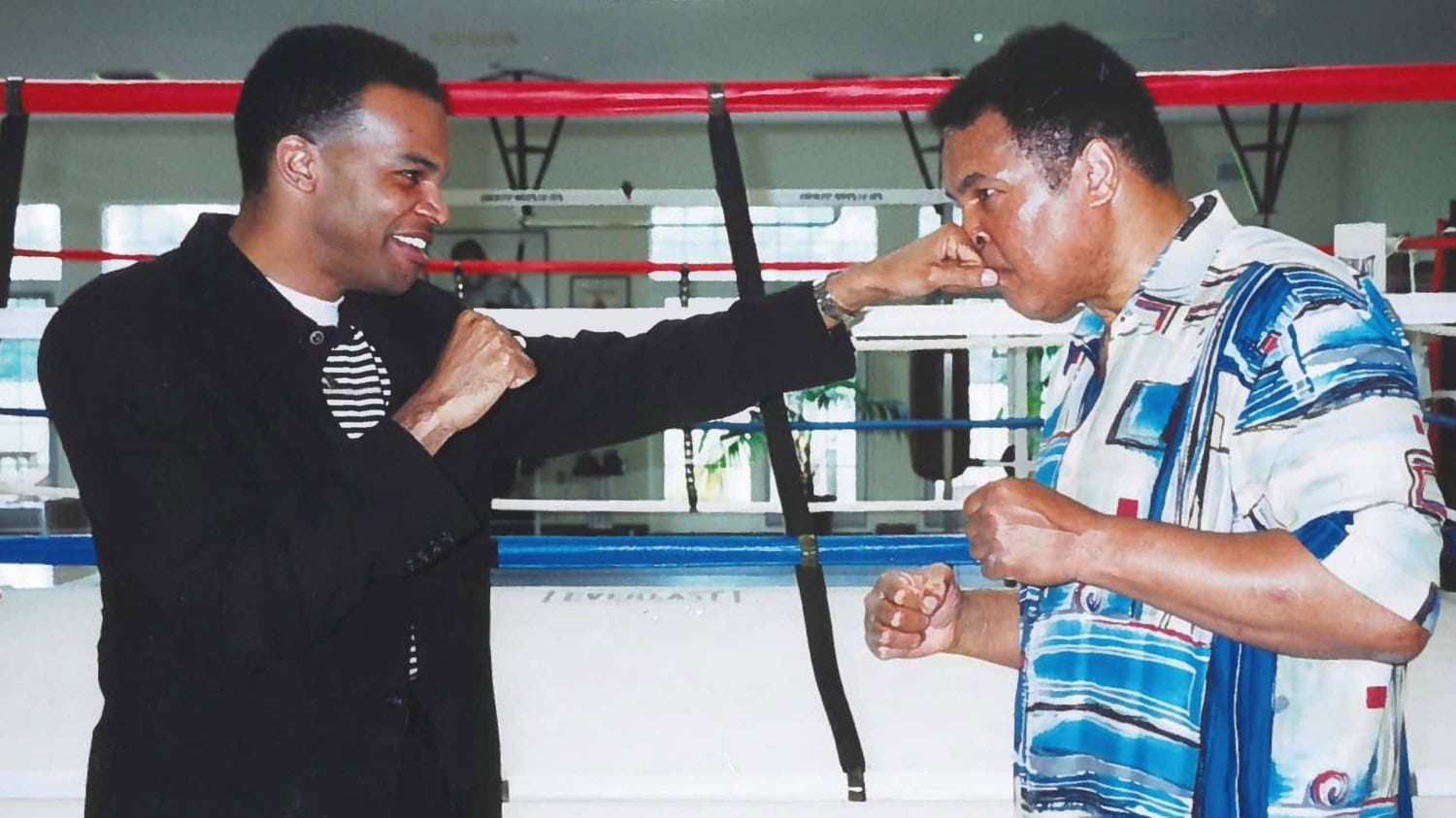 Man wearing black jacket and pants poses with Muhammad Ali outside a boxing ring, with both men in a boxing stance