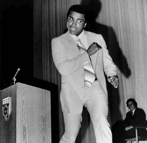 Black and white photo of Muhammad Ali in suit giving boxing pose in front of Harvard University podium