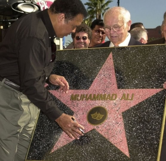 Older man holding up Hollywood Walk of Fame star while Muhammad Ali in button up shirt and gray pants signs with marker