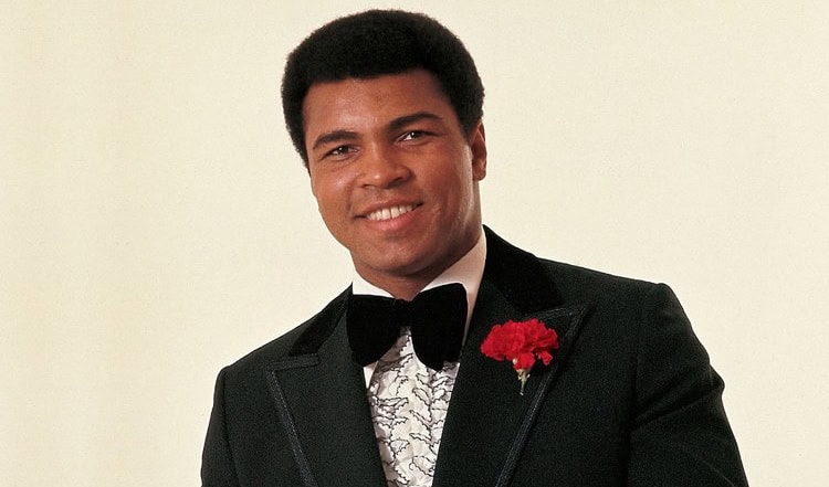 Muhammad Ali posing in black tuxedo with black bow tie smiling at camera