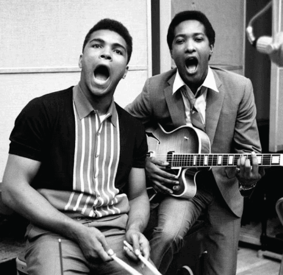 Muhammad Ali sitting at drum set with drumsticks sitting next to Sam Cooke in suit holding a guitar