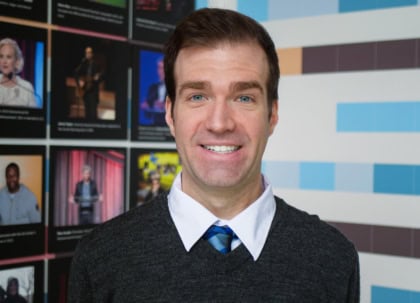 Headshot of man with short hair and blue eyes wearing gray seater and blue tie smiling at camera