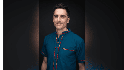 Man with short dark hair wearing blue button down shirt with multi-color edges smiling at camera