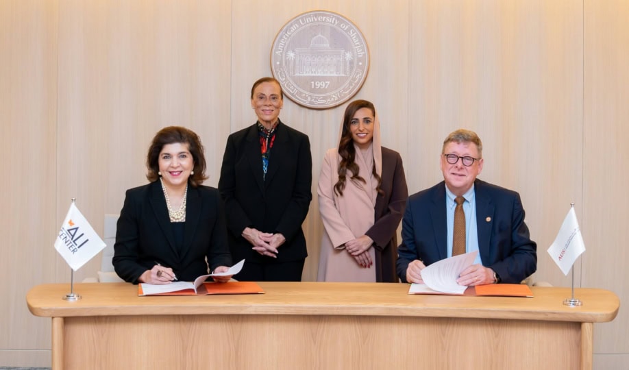 Four people sitting at wooden table and smiling at camera alongside flags with Muhammad Ali Center and American University of Sharjah logos