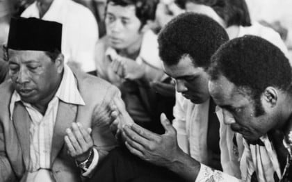 Black and white photo of Muhammad Ali in a room full of men praying