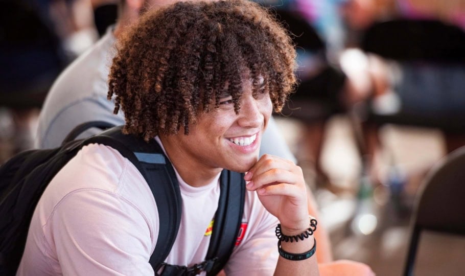 A student wearing a t-shirt and a backpack sits, listens, and smiles